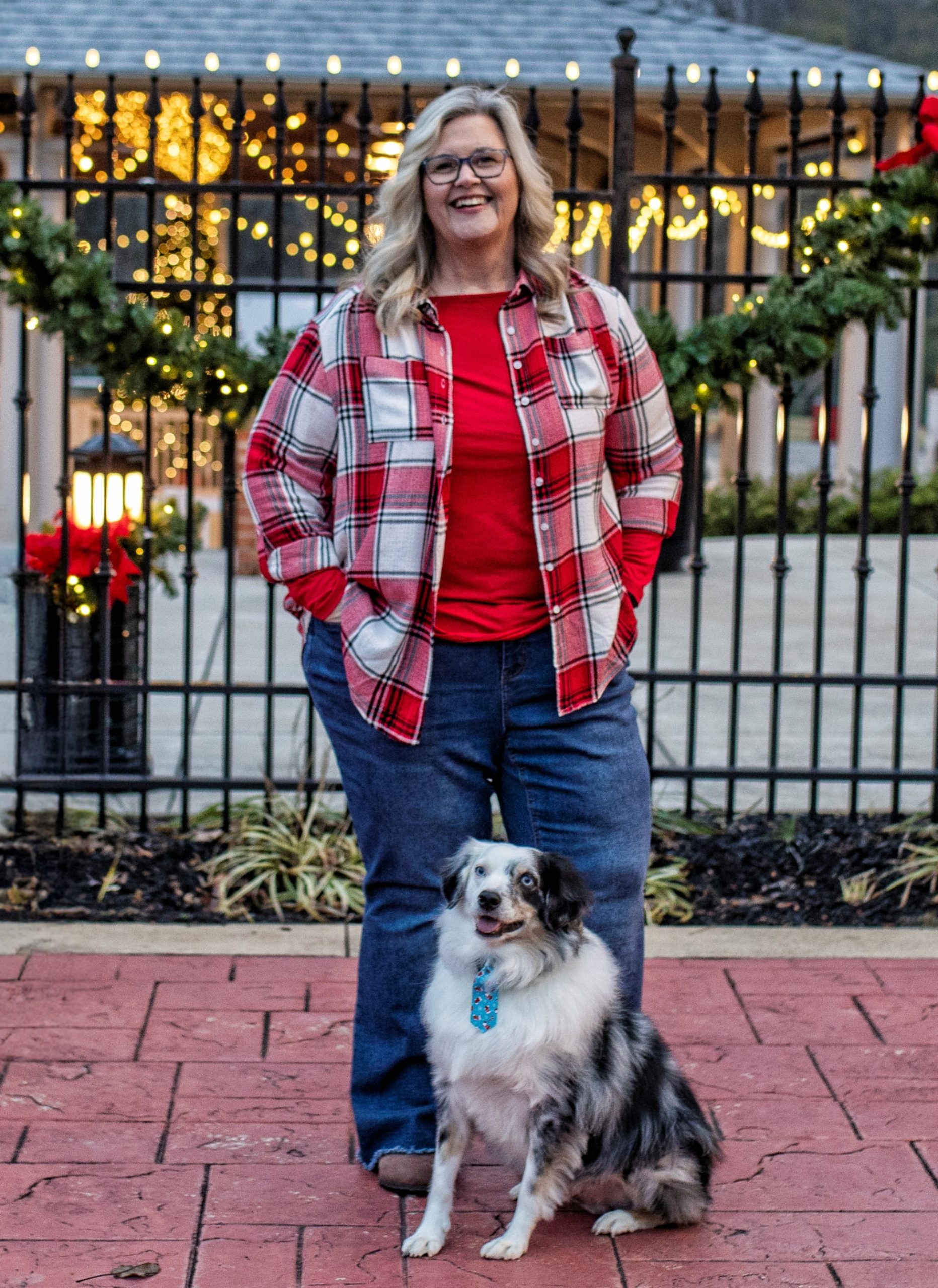 Jennifer Knolton standing outdoors with her dog Teeko during the holiday season, smiling in a casual plaid shirt with festive lights in the background.